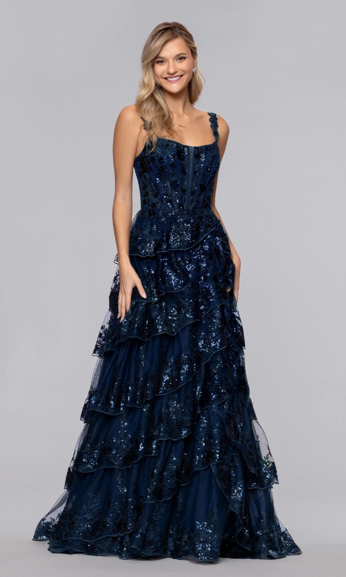 A woman smiles in the Long Prom Dress: Blondie Nites 6126BN, a dark blue, floor-length gown with sequins, a corset bodice, tiered ruffles, and sleeveless design, posing against a plain background.